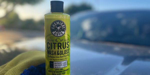 A bottle of premium citrus wash and gloss resting on a microfiber towel beside a car at golden hour